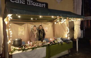 Zwei junge Frauen stehen in einer beleuchteten Weihnachtsbude vor dem Cafe Mozart.