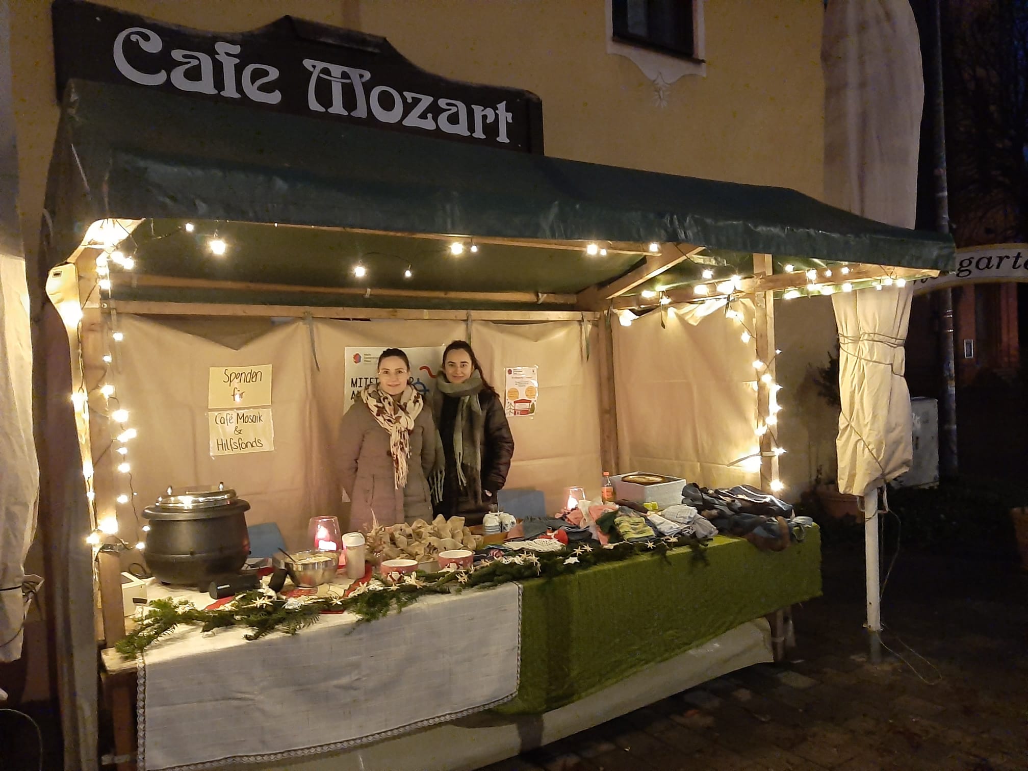 Zwei junge Frauen stehen in einer beleuchteten Weihnachtsbude vor dem Cafe Mozart.